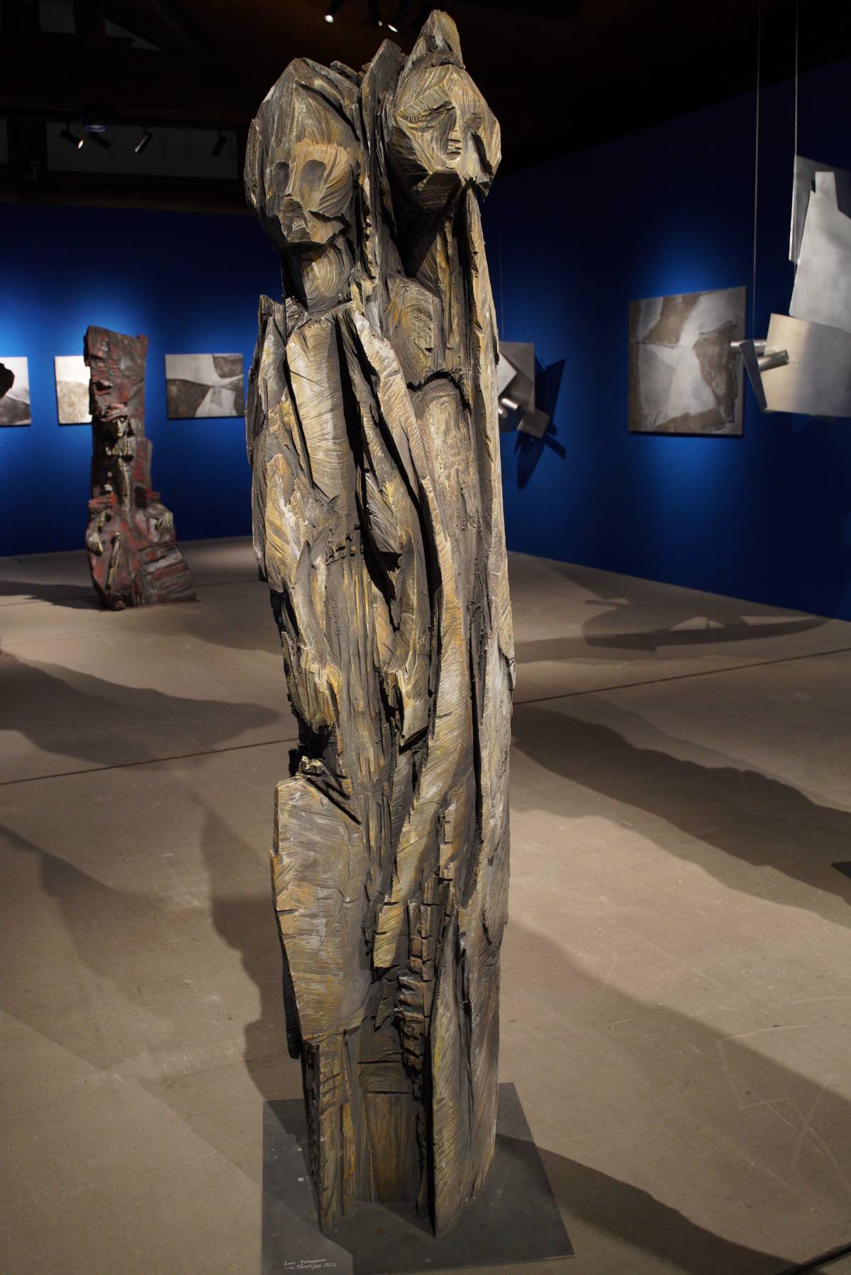 schlanke, lebensgroße Holzfigur auf Metallplatte mit zwei abstrakten Köpfen, sichtbaren Kettensägespuren und rauer, zerklüfteter Oberfläche in Grau- und Ockertönen, vor blauer Wand mit Alubildern