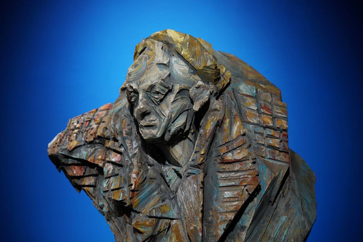 Detailaufnahme einer bunten Holzfigur mit Männerkopf, gelben Kappe und breitem Schal, tiefen Schnittspuren vor blauem Hintergrund