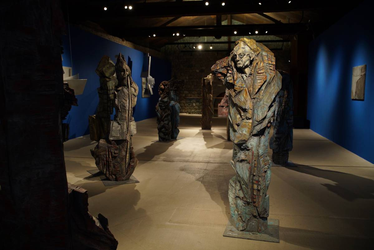 Beleuchtete lebensgroße Holzfigur mit gelber Kappe und sichtbaren Kettensägespuren, umgeben von weiteren Holzfiguren